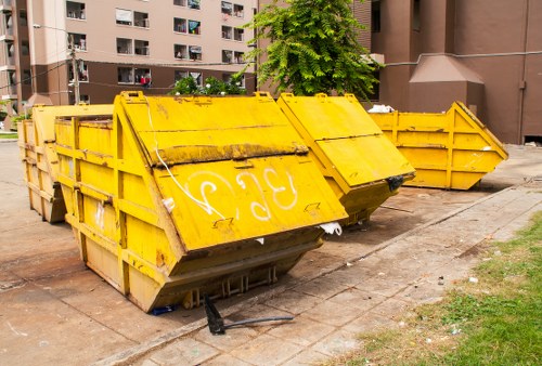 A row of large, heavy-duty metal skip containers painted in bright yellow, positioned on a paved area outdoors in front of a residential apartment complex. The skips are open-topped, with some showing signs of weathering and minor rust, and are arranged in a slight staggered formation. One skip in the foreground has a partially raised side panel, revealing a dark, empty interior. The surrounding environment includes a concrete pavement, a small grassy patch to the right, and a backdrop of multi-storey brown residential buildings with balconies and windows. A leafy green tree is visible behind the skips, adding contrast to the scene. The overall scene depicts commercial waste disposal specifically related to rubbish removal services, with the company's name, Office Clearance Purley, associated with local rubbish collection and clearance activities in the Purley area of London.