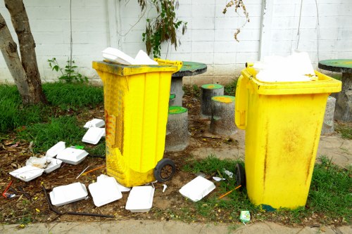 Two bright yellow plastic wheelie bins with closed lids are positioned on a pavement outside a white brick wall, surrounded by scattered white plastic food containers and packaging. The containers vary in size, some are broken or overturned, and debris is strewn across the ground near the bins. Behind the bins, there are small green plants growing along the wall and a tree trunk visible on the left side, suggesting a shared residential or commercial outdoor area in Purley. The scene appears to be part of a rubbish collection or waste disposal context, with the containers used for general waste or food packaging. The lighting indicates daytime with natural sunlight illuminating the area, emphasizing the vibrant colour of the bins and the contrast of the scattered waste against the pavement surface. This image exemplifies typical rubbish removal challenges encountered in local waste management services such as those offered by Office Clearance Purley.