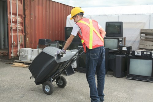 A worker wearing a yellow safety helmet and a high-visibility vest is in an outdoor area with a concrete surface, pushing a large, dark grey plastic rubbish bin on wheels. The background features a red shipping container, several old electronic devices including televisions, a stereo system, and VCRs arranged on a wooden pallet and a stack of wooden pallets on the right side. The setting appears to be a yard or storage area for waste or electronic disposal, with natural daylight illuminating the scene. This image is associated with rubbish removal services provided by Office Clearance Purley, serving the local Purley area. The environment suggests a professional waste collection process, likely involving the disposal of electronic waste or general rubbish collected from commercial or domestic premises in or around Purley postcode area.