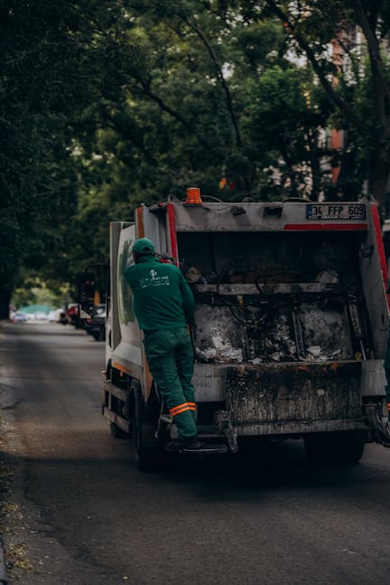 A middle-aged woman with dark hair, dressed in a black short-sleeved shirt and dark trousers, is seen bending over a metal public waste bin to dispose of rubbish. She is holding a large white plastic