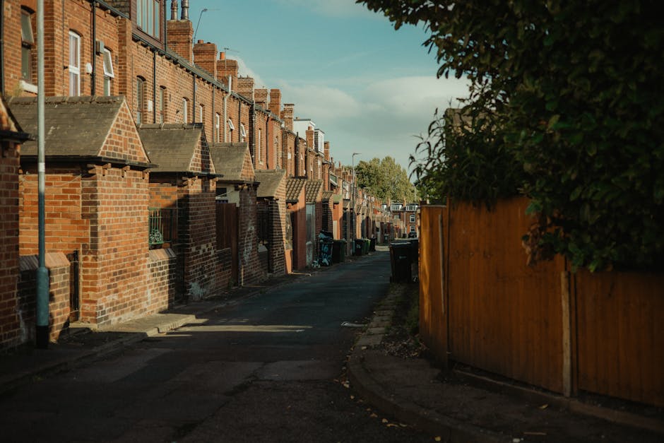 A narrow residential street in a suburban area featuring a row of brick terraced houses with pitched roofs and small front yards. The houses are constructed from red brick with some darker accents, an